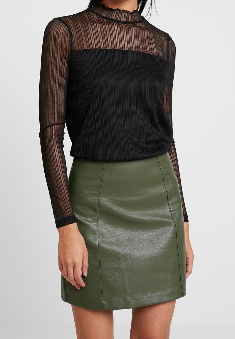 Femme portant un haut transparent noir à manches longues avec des rayures verticales et une mini-jupe en cuir vert olive, debout devant un fond uni.