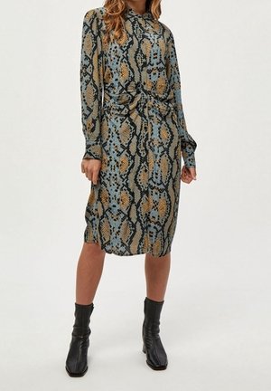 Femme portant une robe boutonnée à manches longues, longueur genou, avec un imprimé serpent bleu et marron, associée à des bottes noires mi-mollet.