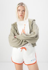 Sudadera con cremallera de color verde claro sobre una camiseta blanca con un gráfico naranja. Pantalones cortos blancos con ribetes naranjas y detalles estampados, con cordón.