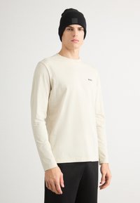 Cămașă crem cu mânecă lungă, guler rotund și un mic logo negru, purtată cu o căciulă neagră strânsă. Design simplu și casual.