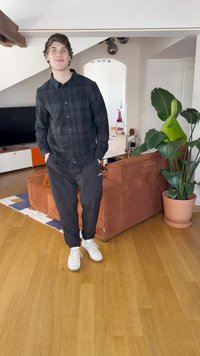 Cămașă cu carouri negre, pantaloni negri texturi și adidași albi. Fundalul include o canapea maro cu sfoară, plante și podea din lemn.