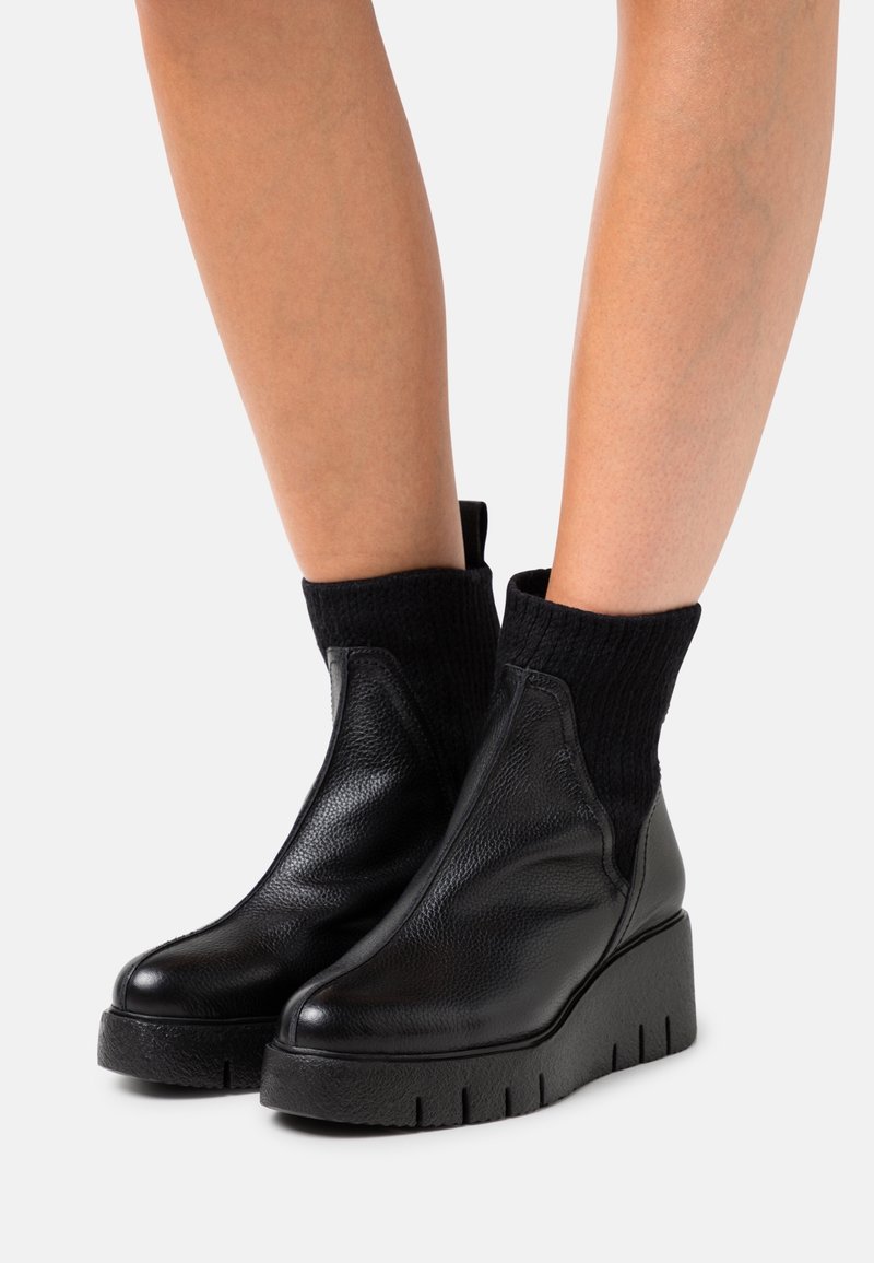 Jambes portant des bottines en cuir noir avec des semelles épaisses et des parties supérieures en maille côtelée semblables à des chaussettes sur un fond uni.