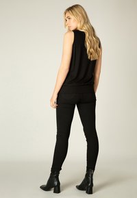 Zwarte mouwloze top en zwarte skinny jeans. Het model staat met de rug naar de camera, waardoor het eenvoudige ontwerp van de top en de strakke pasvorm van de jeans goed zichtbaar zijn. Zwarte hoge laarzen.