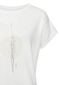 Valkoinen puuvillapaita, jossa on pyöreä pääntie, lyhyet hihat ja abstrakti printti, jossa on hopeanvärisiä yksityiskohtia sekä vaaleanbeige ympyrä.