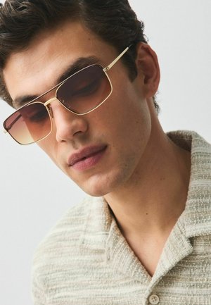 Noormees, kes kannab kuldse raami ja ümmarguste gradientidega päikeseprille ning beeži tekstuuriga kraega särk, vaatab alla heleda tausta ees.