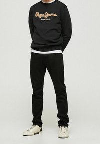 Sudadera negra con el logotipo bordado "Pepe Jeans London" en beige, combinada con pantalones negros y zapatillas de color claro. Diseño simple y casual.