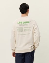 Beige színű pulóver, zöld felirattal a hátsó részén. Az anyaga lágyan hófehér, bordázott mandzsettákkal és alja. A felirat tartalmazza a márkanevet és egy design koncepciót.