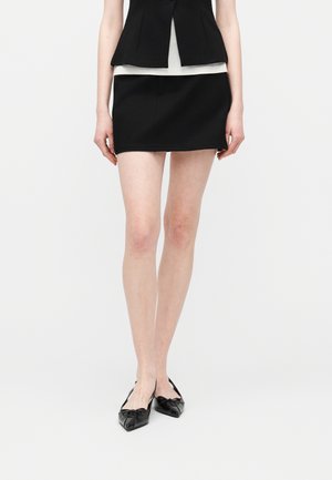 Femme portant une mini-jupe noire, un blazer sans manches noir, un haut blanc et des ballerines pointues noires à nœuds, debout devant un fond blanc uni.