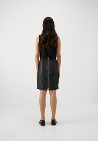 Vestido preto com um corpete ajustado e uma saia de couro, apresentando um fecho central nas costas e uma textura suave. Usado com sapatos pretos.