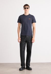 Tricou bleu marin cu mânecă scurtă și un logo mic, asortat cu blugi din denim închis și pantofi negri cu talpă groasă. Modelul poartă ochelari de soare întunecați.