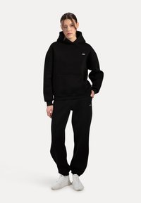 Sort hættetrøje med kængurulomme, prydet med et lille logo. Sammen med matchende sorte joggerbukser og hvide sneakers. Glat tekstur.