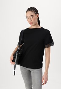 Svart topp med korte ermer, dekorert med blondedetaljer på ermene. Glatt stoff med en klassisk snitt, kombinert med grå jeans og en svart veske.