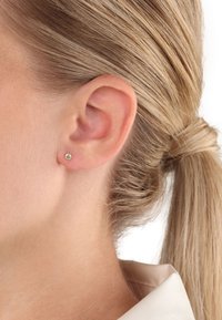 Guldstickare med en liten rund ädelsten, placerad i örsnibben på en person. Håret är stylat bakåt, vilket avslöjar örat.