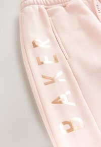 Pantalon de survêtement rose clair avec des lettres dorées métalliques brillantes formant "BAYER" le long de la jambe et une taille élastique.