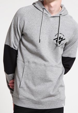 Hombre con sudadera con capucha gris clara, parches negros en los codos y un pequeño logo negro en el pecho izquierdo, con las manos detrás de la espalda.