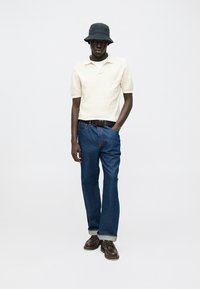 Κρεμ πόλο μπλούζα με κοντά μανίκια, μπλε τζιν παντελόνι, μαύρο καπέλο τύπου bucket, σκούρα καφέ παπούτσια, με κλασική εφαρμογή και ανασηκωμένα ρεβέρ στο τζιν.