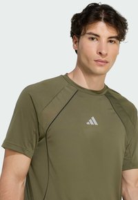 Camisa deportiva de color verde oliva con mangas cortas, que presenta un cuello redondo y acentos negros, exhibida con un pequeño logo blanco en el pecho.