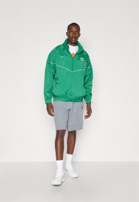 Grøn Nike vindjakke med lynlås krave, hvide detaljer og logo, båret over en orange skjorte og grå shorts, parret med hvide sneakers.