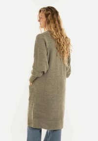 Vrouw met lang golvend haar in een lange olijfkleurige gebreide cardigan met zakken en blauwe jeans, staand met haar gezicht naar links tegen een witte achtergrond.