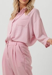 Camicia button-up di color rosa chiaro con tessuto testurizzato, una sola tasca sul petto e maniche ampie, abbinata a pantaloni a vita alta coordinati.