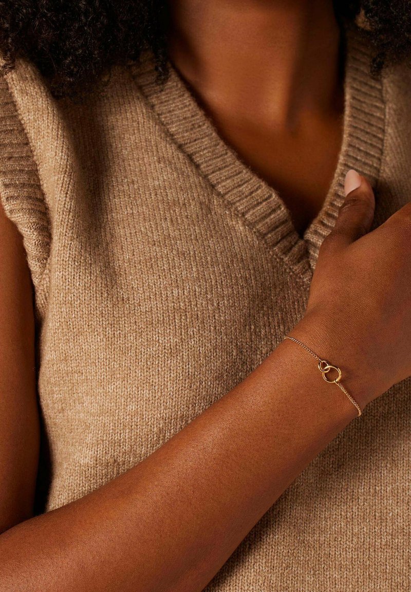 Gouden kettingarmband met met elkaar verweven cirkelvormige accenten, gedragen op een donkere huidtonen. Grijs-bruin gebreid mouwloos trui op de achtergrond.
