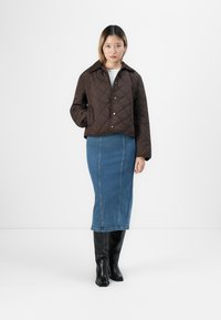Mujer vestida con una chaqueta acolchada marrón, una falda midi de mezclilla azul y botas altas negras, de pie frente a un fondo blanco.