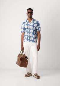 Korte blauwe shirt met wit bladerenpatroon, witte broek, bruinleren tas en beige sandalen. Model draagt donkere zonnebril.