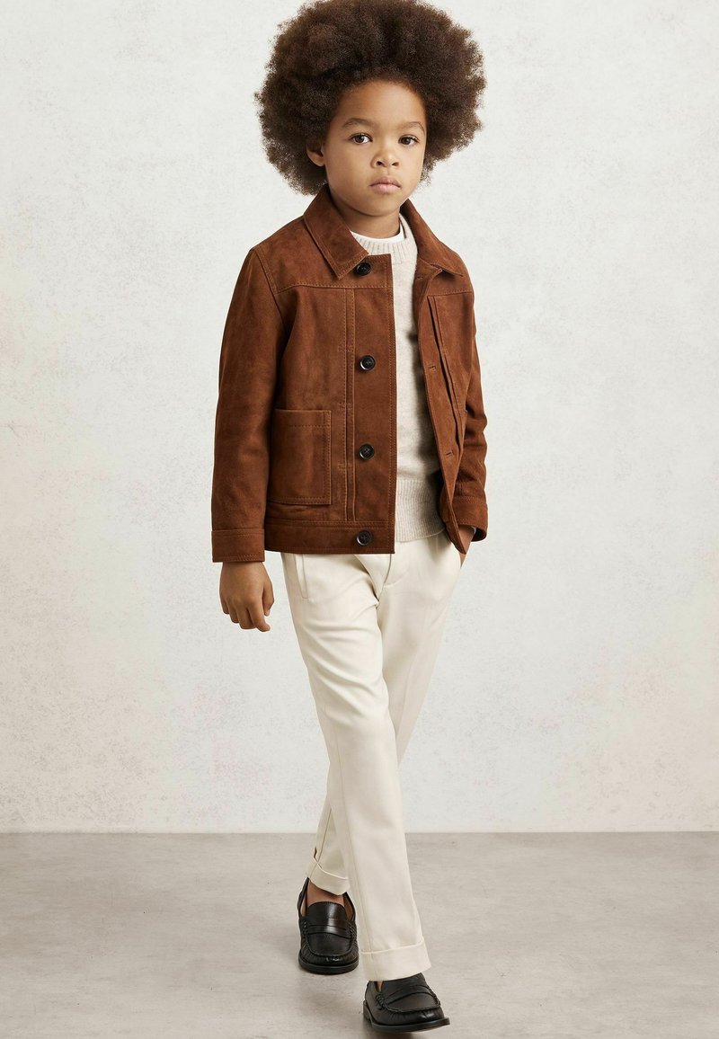 Brun mockajacka med krage, knappstängning och sidofickor, tillsammans med krämfärgade byxor och svarta slip-on skor.