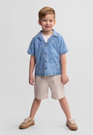 Uśmiechnięty chłopiec stojący z rozstawionymi nogami, ubrany w niebieską koszulkę z krótkim rękawem z nadrukiem palm, beżowe spodenki i beżowe trampki.