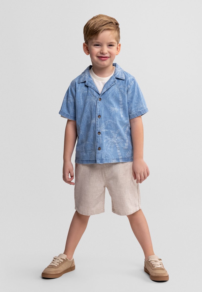 Garçon souriant debout, jambes écartées, portant une chemise à manches courtes bleue avec un motif de palmier, un short beige et des baskets beiges.