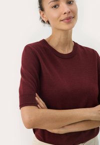 Maglione corto a maniche corte color bordeaux con collo tondo, caratterizzato da una texture liscia e polsi a coste. Il modello è in piedi con le braccia incrociate.