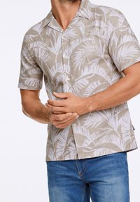 Chemise à manches courtes avec col, de couleur beige clair avec un motif de feuilles tropicales blanches. Le tissu semble respirant ; boutons sur le devant.