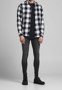 Hombre vistiendo una camisa de cuadros en blanco y negro sobre una camiseta negra, jeans ajustados oscuros, zapatos negros y anillos en los dedos, de pie sobre un fondo gris.