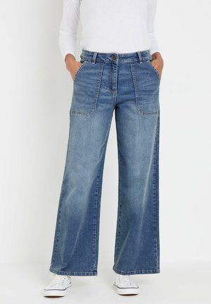 Donna che indossa jeans di jeans blu a vita alta con gamba larga e grandi tasche frontali, abbinati a sneakers bianche e una maglia a maniche lunghe bianca.