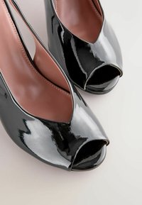 Czarne lakierowane szpilki peep-toe o gładkim wykończeniu i zakrzywionym wzorze wycięcia, z cielistym wnętrzem i wysokim obcasem w kształcie szpilki.