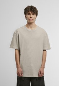 T-shirt bej din bumbac cu o croială lejeră, guler rotund și mâneci scurte. Prezintă un material texturat. Modelul poartă pantaloni scurți închiși la culoare.