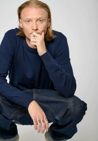 Pull en tricot bleu marine avec des manches texturées, associé à un jean foncé ample. Une main présente un motif de tatouage noir sur les doigts. Arrière-plan neutre.