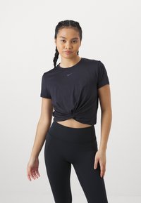 Fekete, rövid ujjú sportpóló, elöl csomózott dizájnnal és egy kis logóval. Fekete, magas derekú leggings-szel viselve.