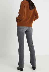 Suéter marrón de punto con un corte relajado, combinado con jeans grises acampanados. El conjunto presenta costuras sutiles y calzado negro con tachuelas.
