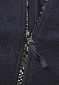 Close-up van een gedeeltelijk dichtgestikte donkermarine fleecejack, met zichtbare metalen ritssluitingen en een stoffen trekker aan de rits.