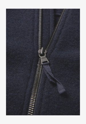 Nahaufnahme einer teilweise zugeknöpften dunkeln Marine-Fleecejacke, die die Metallzähnchen des Reißverschlusses und die Stofflasche am Reißverschluss-Schieber zeigt.