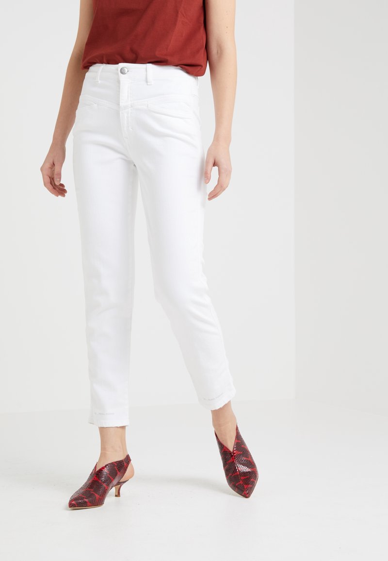 Mujer vistiendo pantalones cortos blancos y zapatos de tacón bajo con punta roja y estampado, de pie contra un fondo blanco liso.