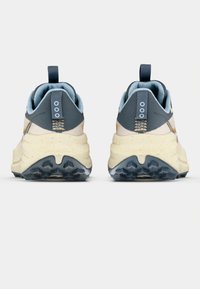 Urheilukenkien takaosa, jossa on beige ja harmaa värimaailma, teksturoidut pinnat ja kuvioitu pohja paremman pidon takaamiseksi. Kenkässä on vetotappi.