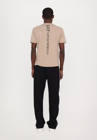 Beige bavlněné tričko s vertikálním černým textem "EA7 EMPORIO ARMANI" na zádech; kombinováno s černými kalhotami s bílými bočními pruhy.