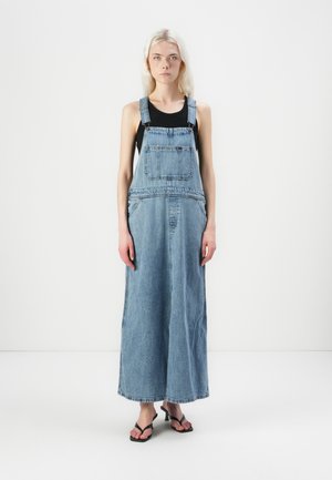 Jeune femme aux longs cheveux blonds portant un haut noir sans manches sous une longue robe salopette en jean et des sandales noires à bout ouvert.