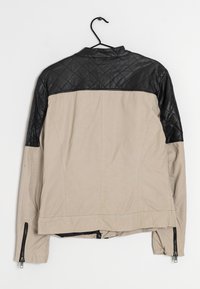 Ljusbeige jacka med svart kviltat läder på axlarna, dragkedjeslut och ståkrage. Materialet är en blandning av tyg och läder.