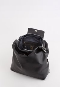 Mochila de cuero negra con un acabado suave y texturizado. El cierre con cremallera revela un interior azul marino con un bolsillo y una etiqueta de marca. Acentos de hardware plateado.