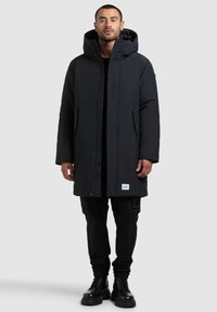 Melnā izolētā parka ar kapuci, taisna griezuma, priekšējā aizdare ar pogām un sānu kabatām. Tekstūrēta auduma ar logotipa detaļu apakšmalā.