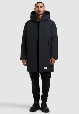 Zwarte geïsoleerde parka met capuchon, met een rechte snit, sluiting met drukknopen aan de voorkant en zijzakken. Gelaagd materiaal met logodetail aan de onderrand.