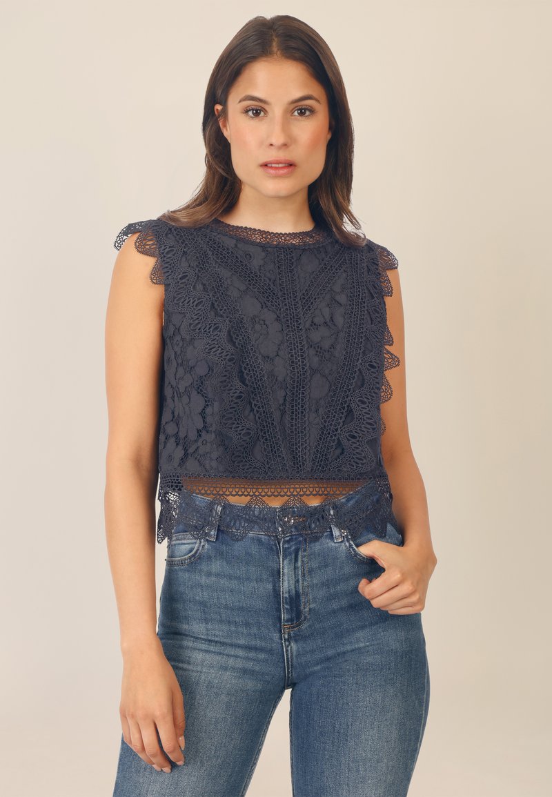 Top corto blu navy con dettagli in pizzo intricati, bordi scallop e un design testurizzato, abbinato a jeans blu aderenti.